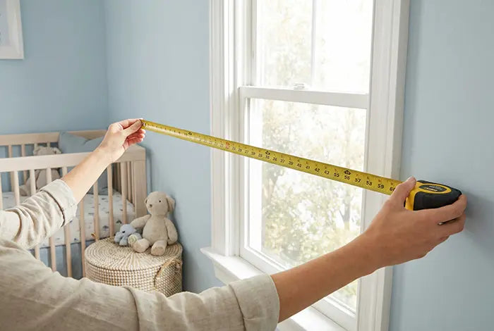 Hands extending a tape measure across a window to measure the full width of the wall for a wallpaper mural.